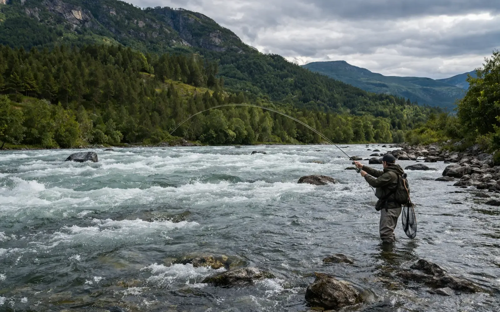 Fluefisker vader i bred lakseelv med strøm og fjell i bakgrunnen